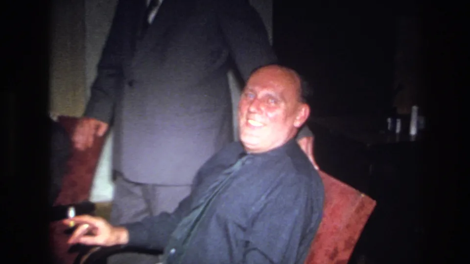 1967 France home-movie frame of a bald man in a dark shirt and tie smiling from a red chair as a suited man stands behind him.