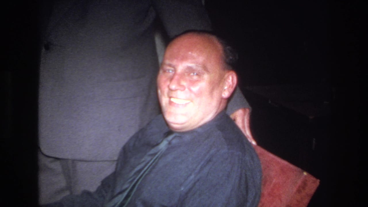 Closer view of the seated man smiling, with a suited figure partly visible behind him.