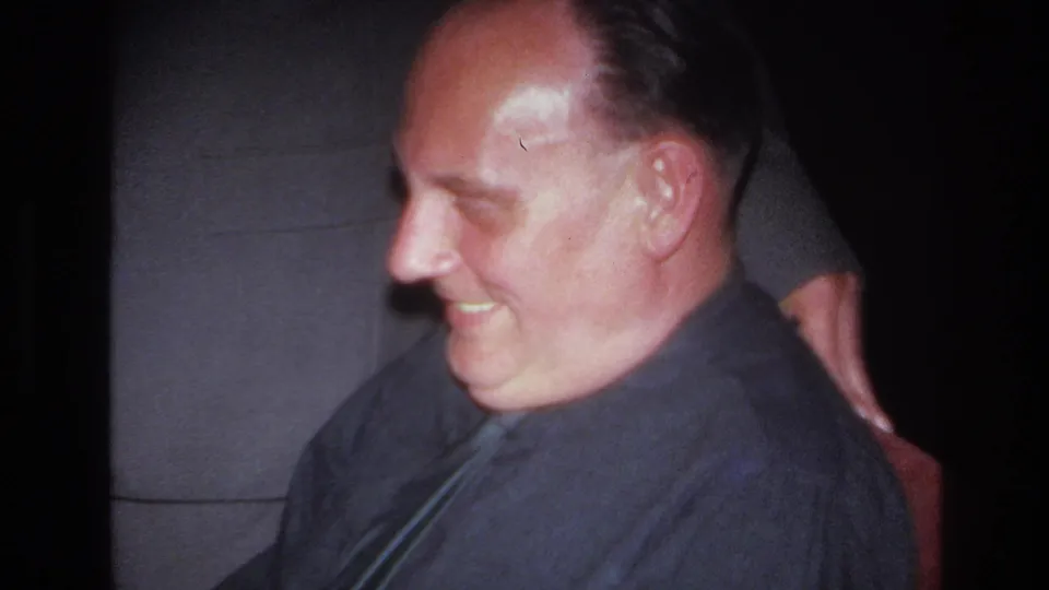 A side-profile view shows the seated man smiling in a dim French home-movie interior, with a suited person close behind him.