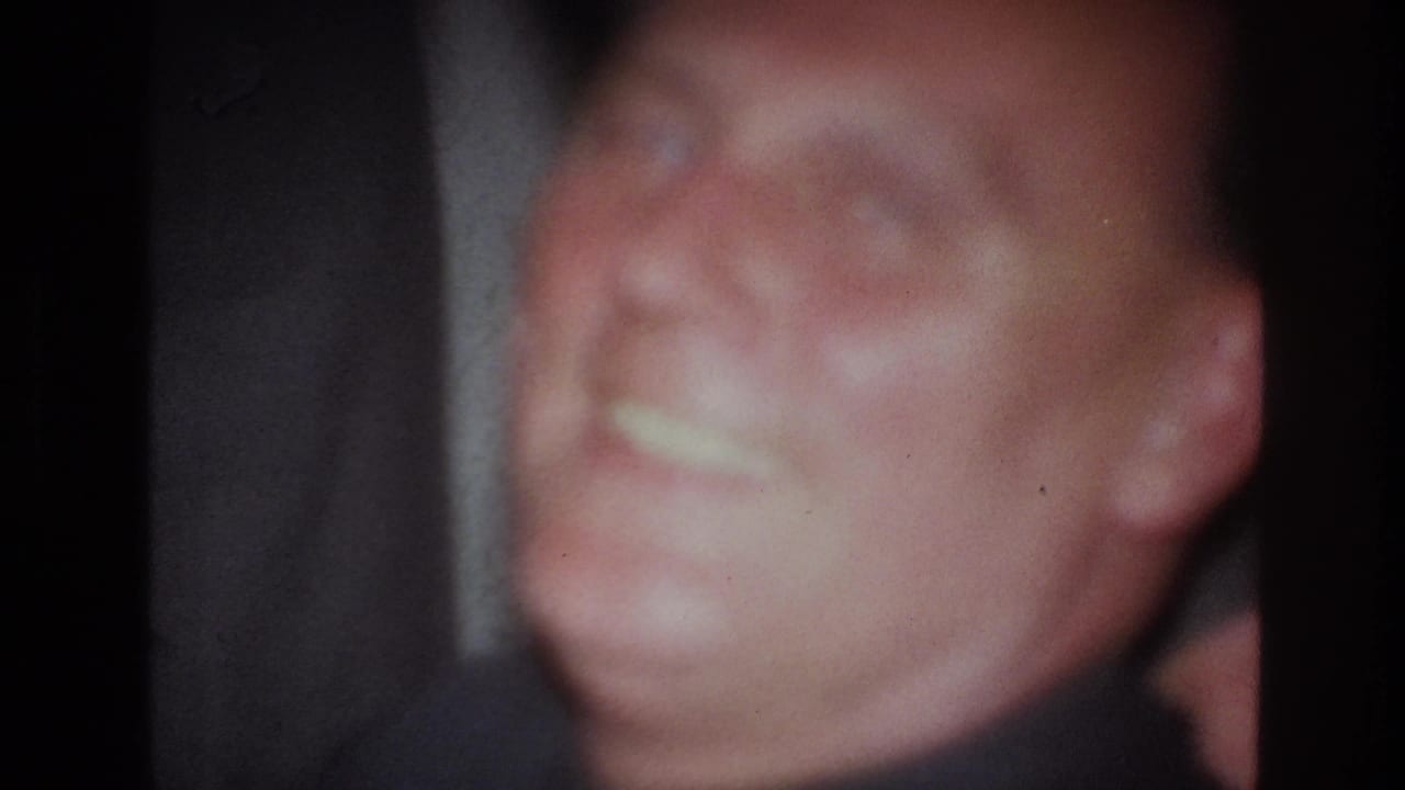 Soft 1967 France 8mm frame showing a seated adult man smiling with a cigarette in a dark indoor setting.