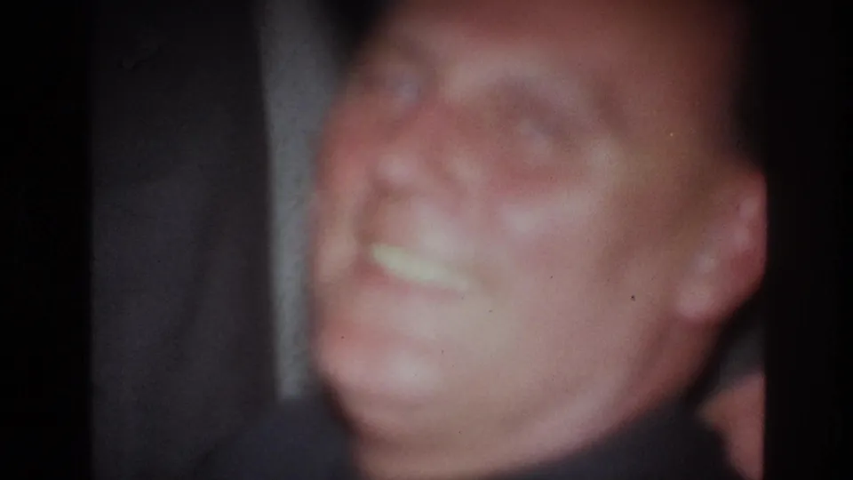 Soft 1967 France 8mm frame showing a seated adult man smiling with a cigarette in a dark indoor setting.