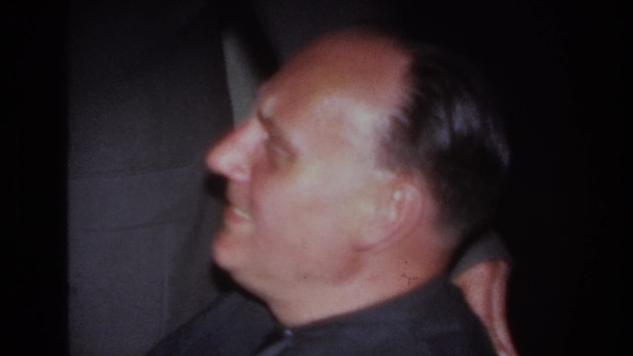 Seated man in profile smiles while another suited man stands behind him.