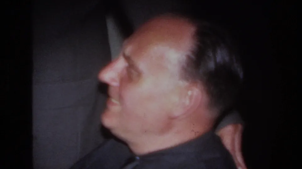 1967 France home-movie frame of a seated man in profile, smiling beside a standing man in a suit indoors.