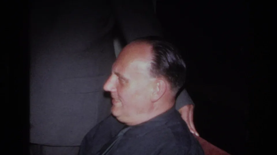 1967 France 8mm still showing the seated man smiling in profile, with a standing suited adult partly visible behind him.