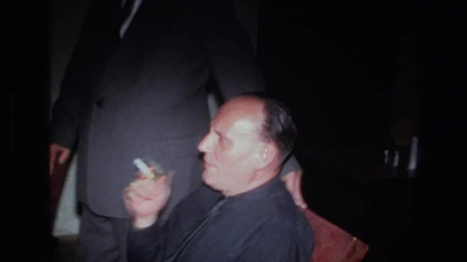 1967 France 8mm frame of a seated man holding a cigarette, with another suited figure partly visible in the dim room.