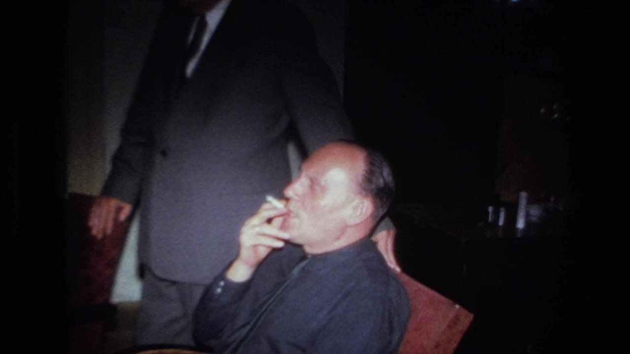 1967 France 8mm frame of a seated adult man smoking in a leather armchair while another suited man stands close by in a dim room.
