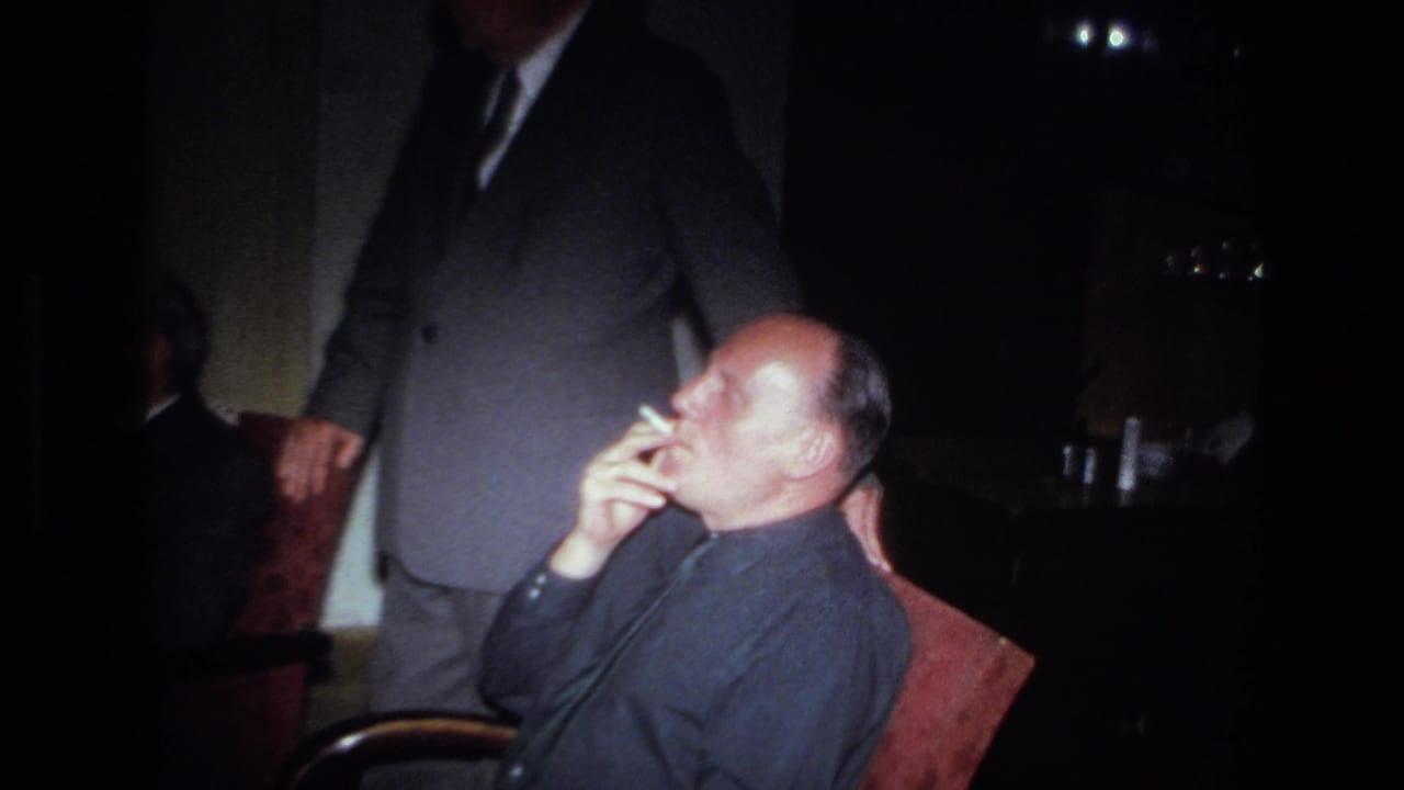 A seated man smokes in profile in a dim 1967 France interior, with a suited adult partly visible behind and beside him.