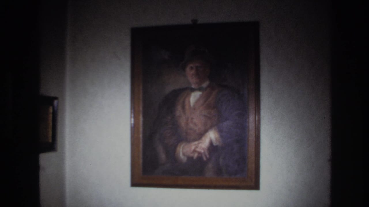 1967 France 8mm frame showing a framed portrait of a man in a hat, vest, and bow tie on a softly lit wall.