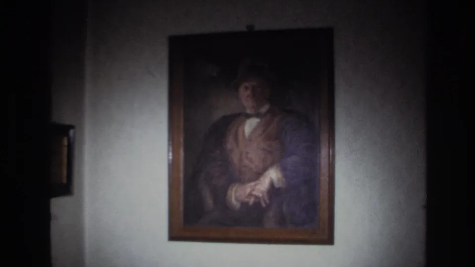 1967 France 8mm frame showing a framed portrait of a man in a hat, vest, and bow tie on a softly lit wall.
