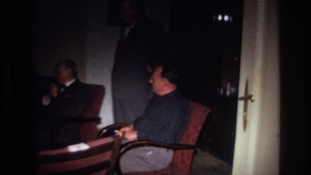 1967 France home-movie frame of men in a low-lit private interior, with one seated in the foreground and another standing behind.