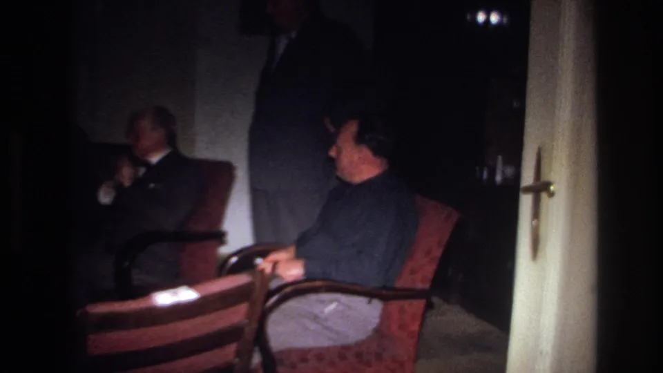 1967 France 8mm frame showing men in formal dress seated in upholstered chairs as another figure stands nearby.