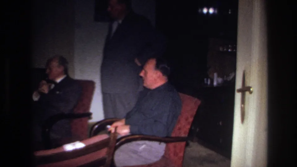 1967 France home-movie frame of a seated man in the foreground, another man behind him, and a standing figure in a private interior.