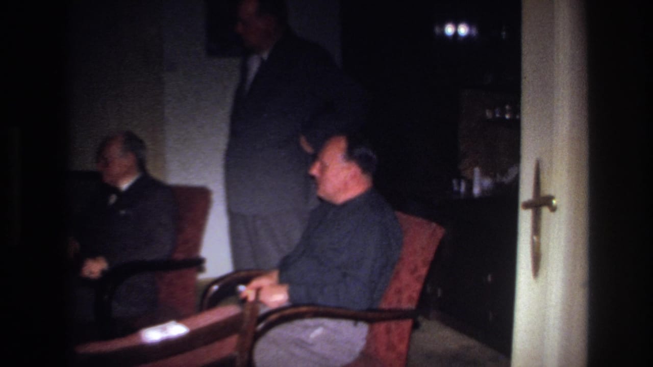 1967 France 8mm frame showing three adult men in suits inside a dark private room, with two seated and one standing.