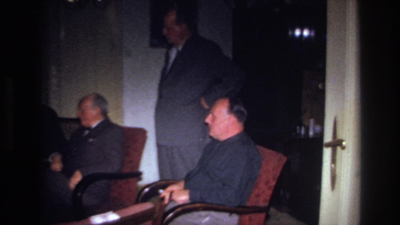 Two seated men and a standing man are framed beside an open doorway in a dark interior.
