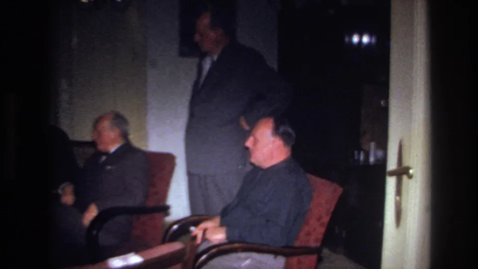 1967 France home-movie frame with suited men gathered near an open door, one seated in the foreground and another partly turned away.