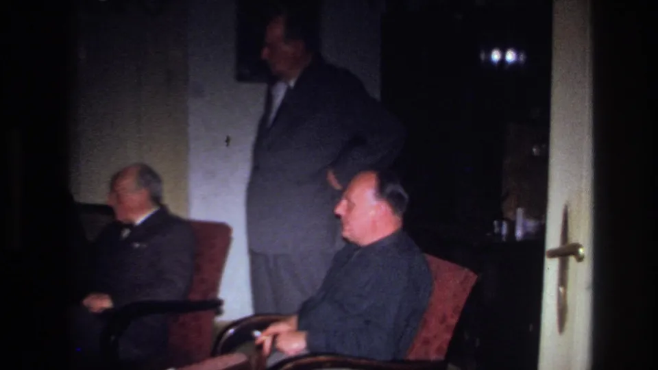 1967 France 8mm frame showing a standing suited man beside two seated men, with armchairs and a shadowed interior visible.