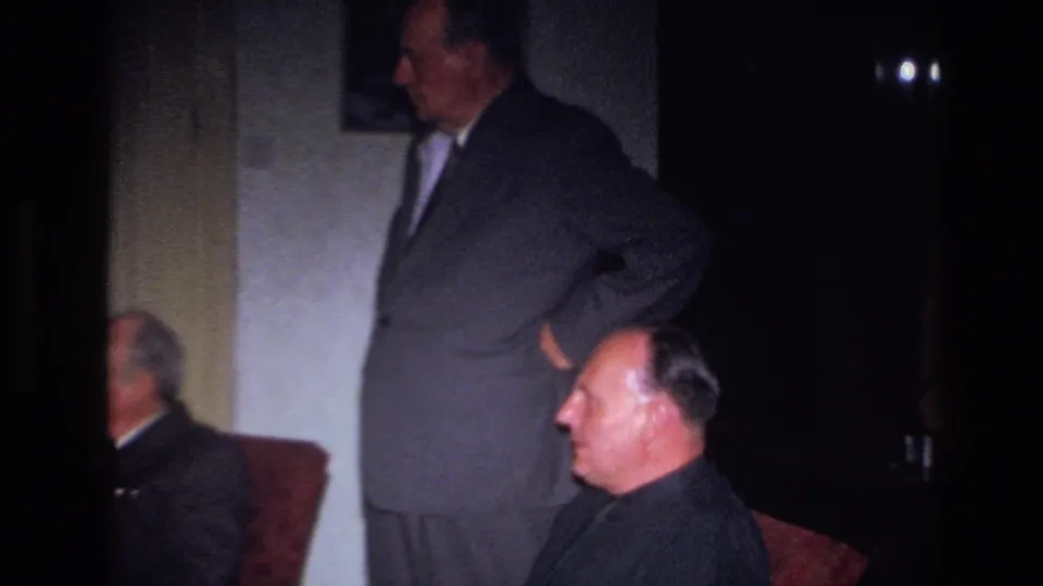 1967 France home-movie frame with a seated man in profile, a standing suited man behind him, and a third seated man partly obscured.