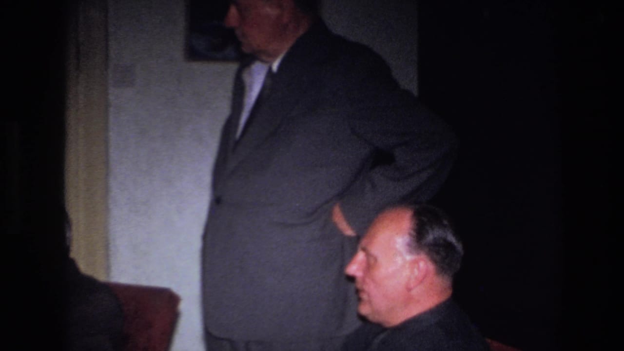 1967 France 8mm frame showing a dim interior with a standing suited man and a seated man in profile near patterned seating.