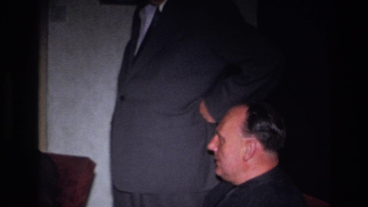 A seated man in a dark suit is shown in profile beside a standing suited figure.