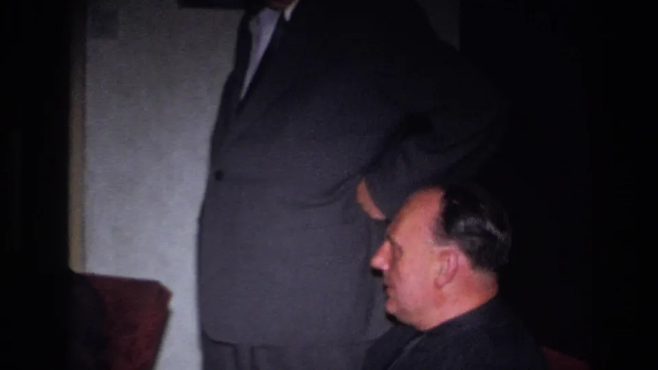The frame shows a formal indoor scene in France, with a seated man in profile and a standing man partly cropped above him.
