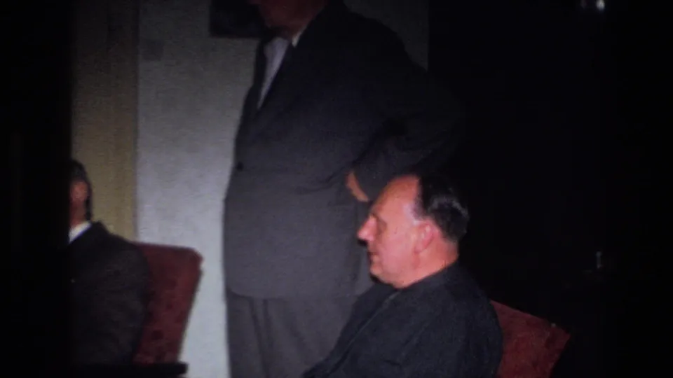 This 1967 France home-movie frame shows a seated man in profile, a cropped standing man, and part of another seated figure at left.
