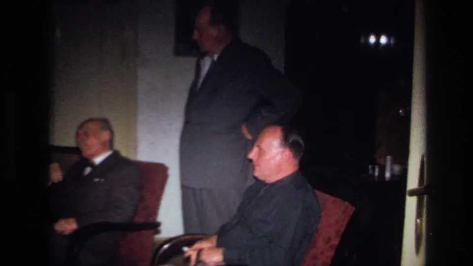 The wider 1967 France frame shows three adult men at a formal indoor gathering, two seated in armchairs and one standing near a doorway.