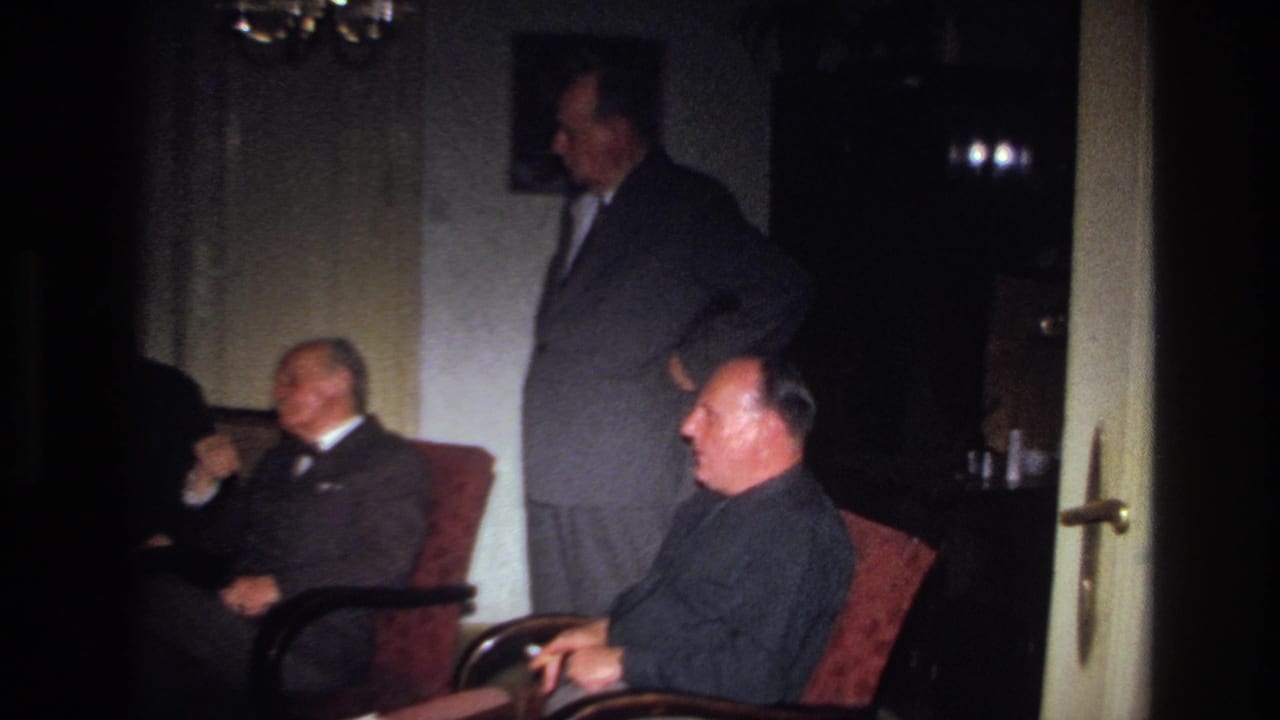 1967 France home-movie frame showing suited adults gathered in a dim private living room with armchairs and wall decor.
