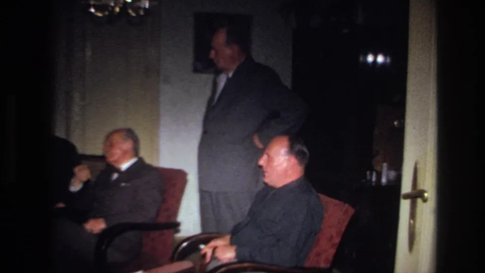 1967 France home-movie frame showing suited adults gathered in a dim private living room with armchairs and wall decor.