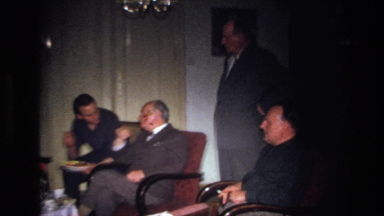 Men in suits talk in armchairs beside a small table in a living room.
