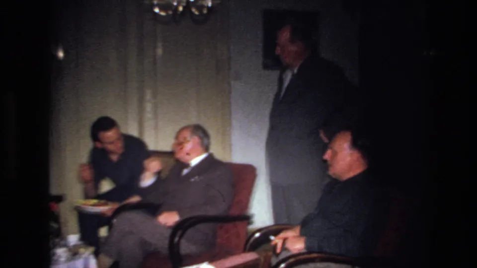 1967 France frame of men in formal attire conversing in armchairs inside a dim living room, with a plate or tray nearby.