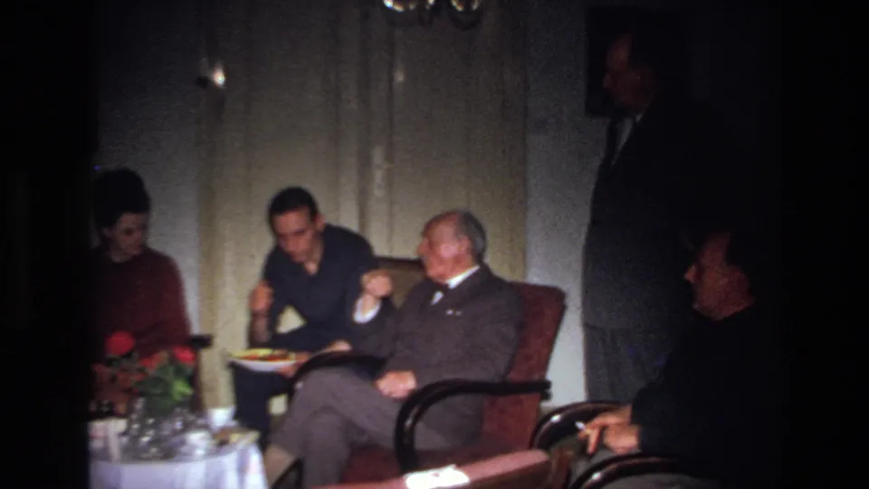 1967 France home-movie frame showing a woman in red and suited men seated near a small table with red roses and food.