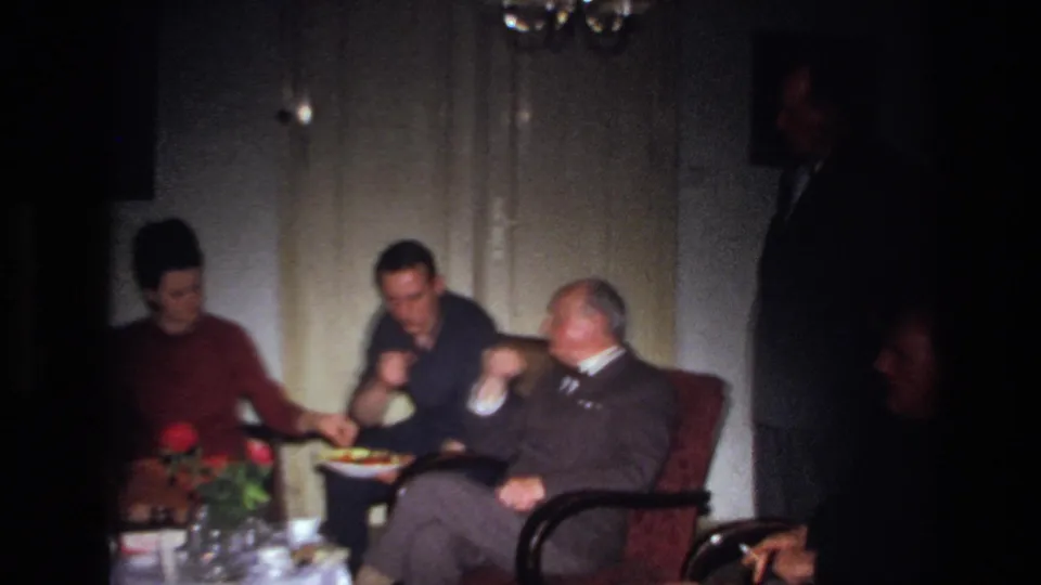 1967 France frame of adults in a private living room, including a woman in red and suited men near red roses and a plate of food.