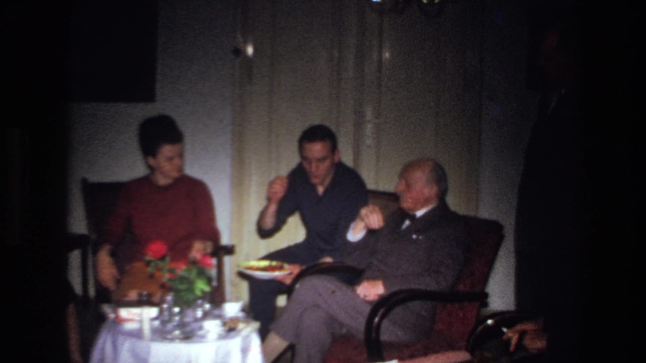 1967 France home-movie frame showing adults in a dim living room, with a younger man serving food beside an older man in a suit.