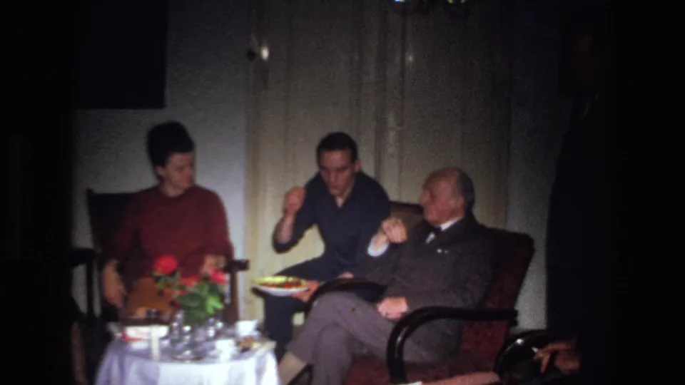 1967 France home-movie frame showing adults in a dim living room, with a younger man serving food beside an older man in a suit.