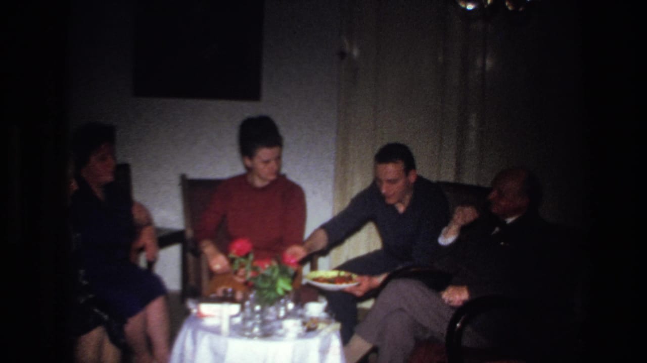 A woman in a red sweater sits at a flowered table as a man offers food to an older seated man.