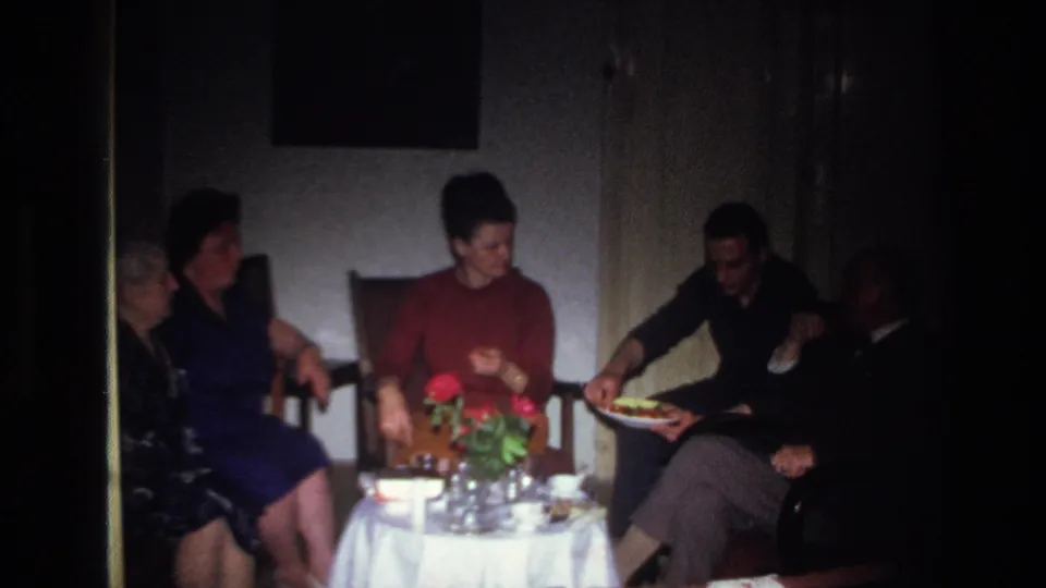Home-movie frame from France in 1967 showing seated adults around a white-covered table with red flowers and food.