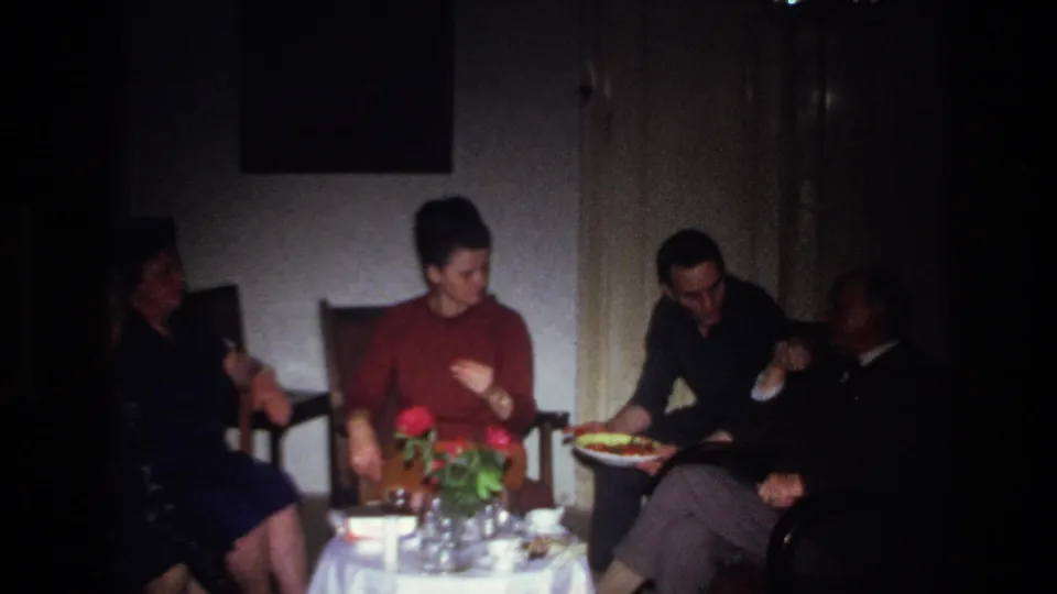 1967 France family gathering frame with adults seated in a private living room beside a small table with flowers and dishes.