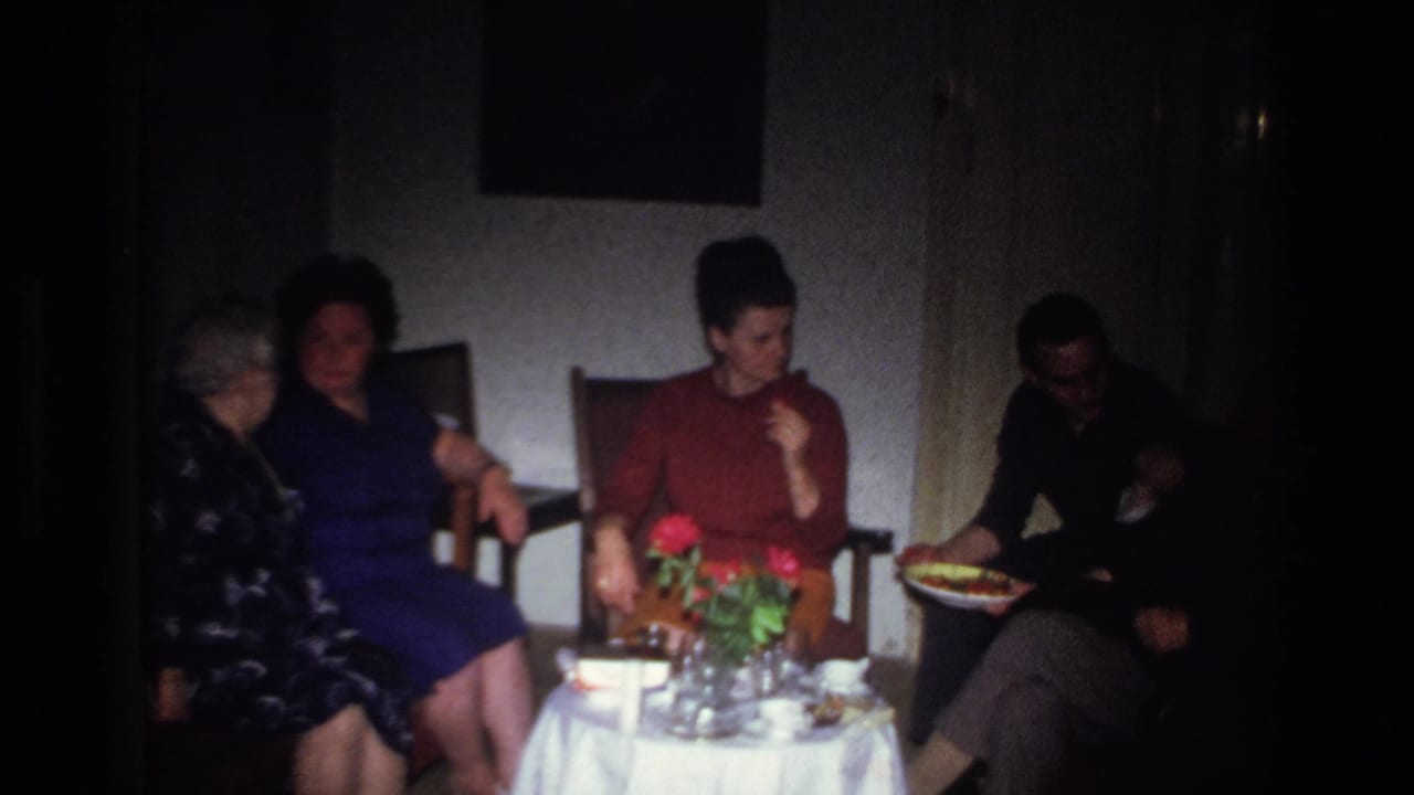 1967 France home-movie frame showing women seated in a private room around a small table with red flowers, glasses, and food.