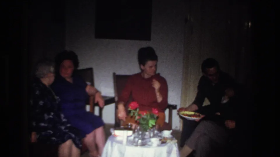 1967 France home-movie frame showing women seated in a private room around a small table with red flowers, glasses, and food.