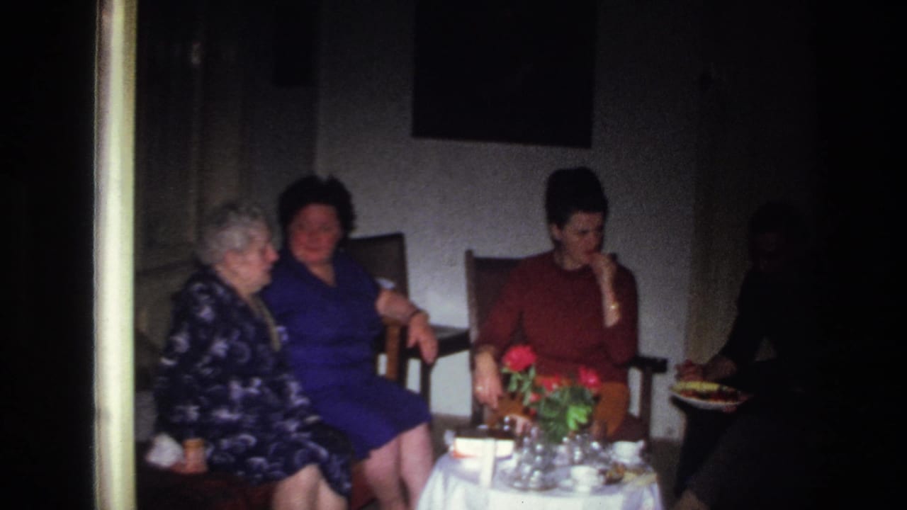 Three women sit beside a table with red flowers while another person is partly visible in shadow.