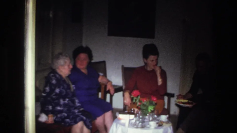 1967 France home-movie frame of women seated near a flower-topped table, with a shadowed person holding food at the edge.