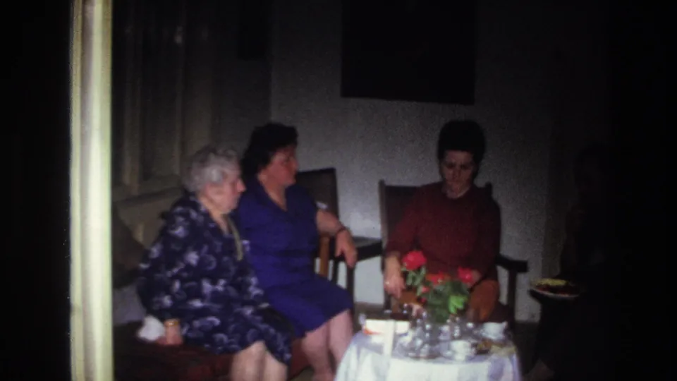 1967 France home-movie frame showing women seated close together in a private interior, beside flowers, dishes, and curtains.