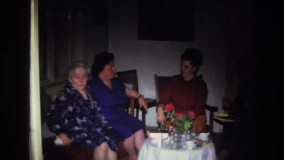 1967 France home-movie frame of three women seated around a small table with red flowers in a dim private room.