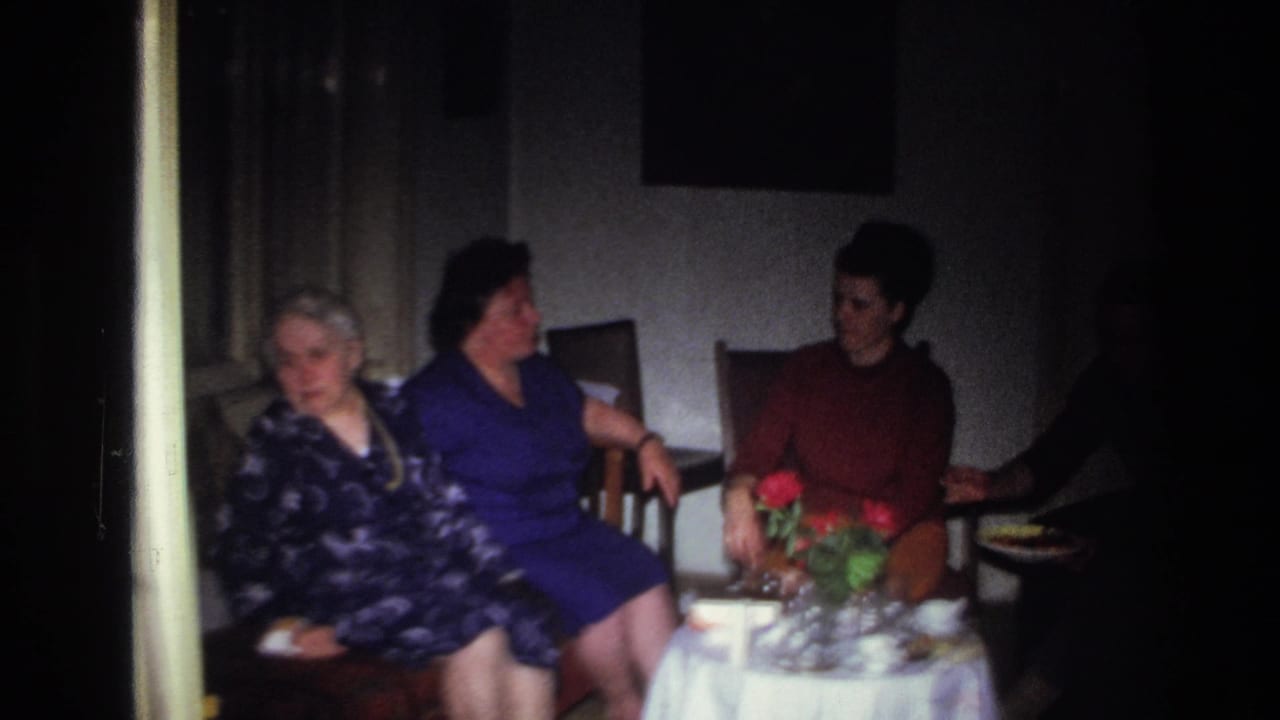 1967 France home-movie frame of three women seated indoors around a small tea table with red flowers and glassware.