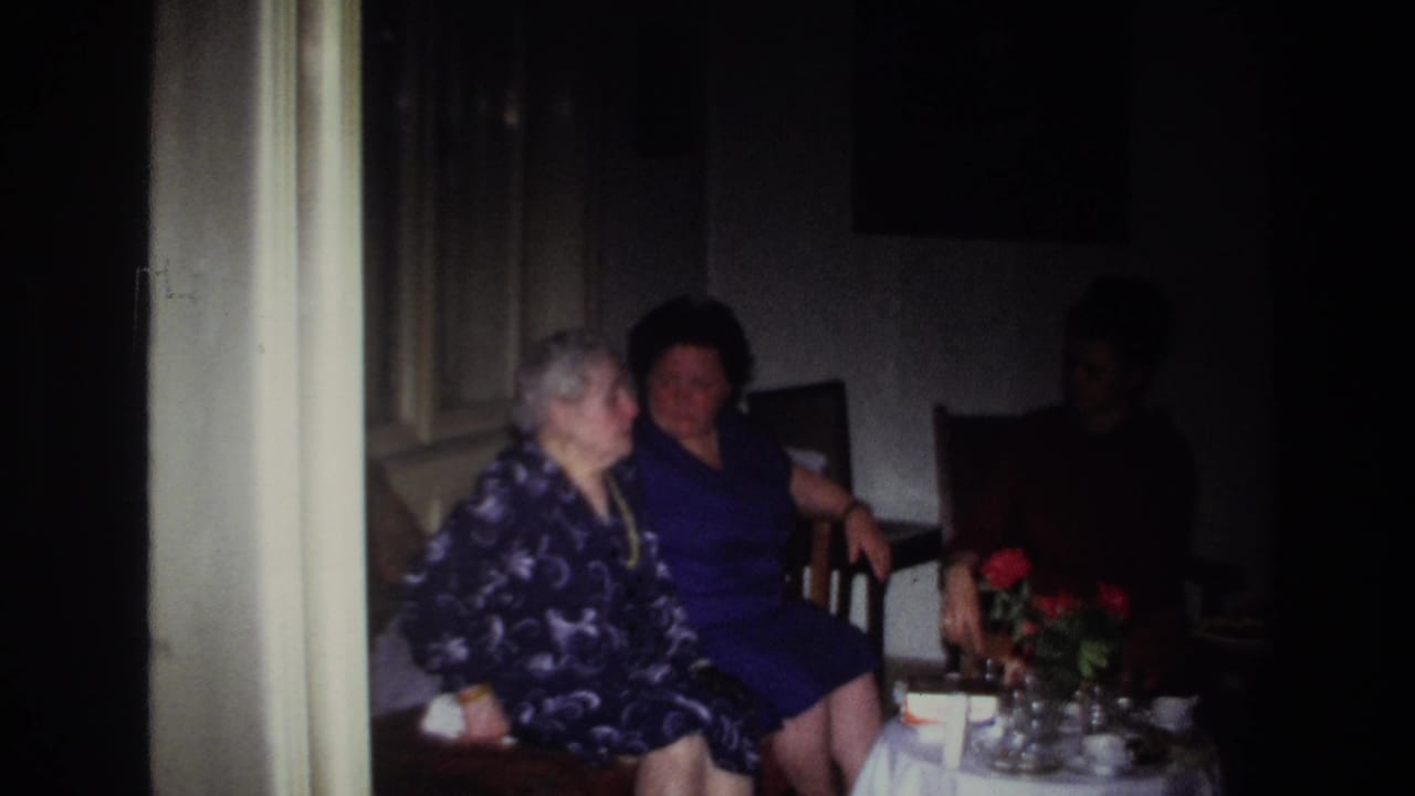 Women seated near a table with red flowers, with part of the room falling into shadow.