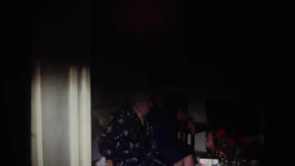 Dark 1967 France home-movie frame with seated women partly obscured, a small table, and red flowers visible in the living room.