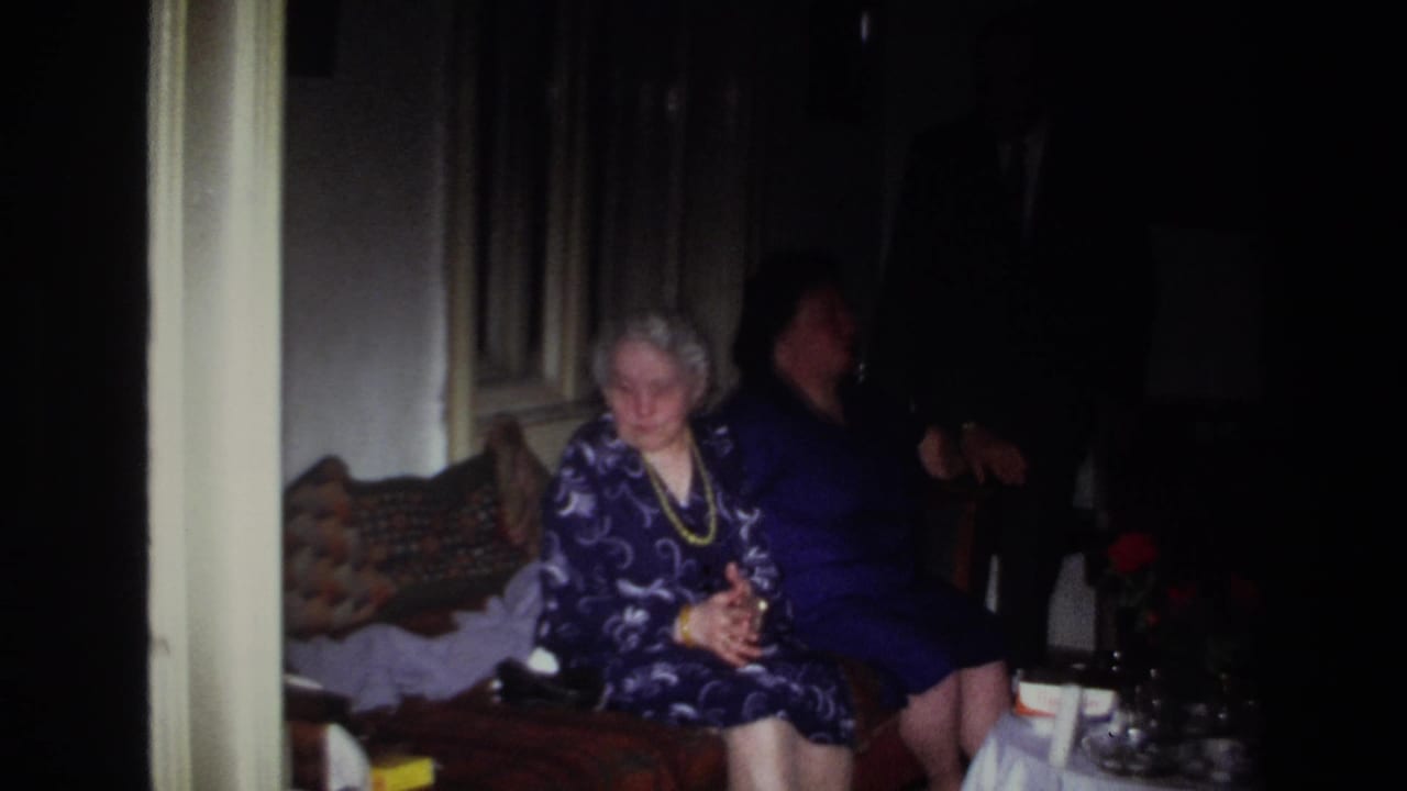 In 1967 France, two women sit close together in a dim private room, with a patterned couch and table partly visible.