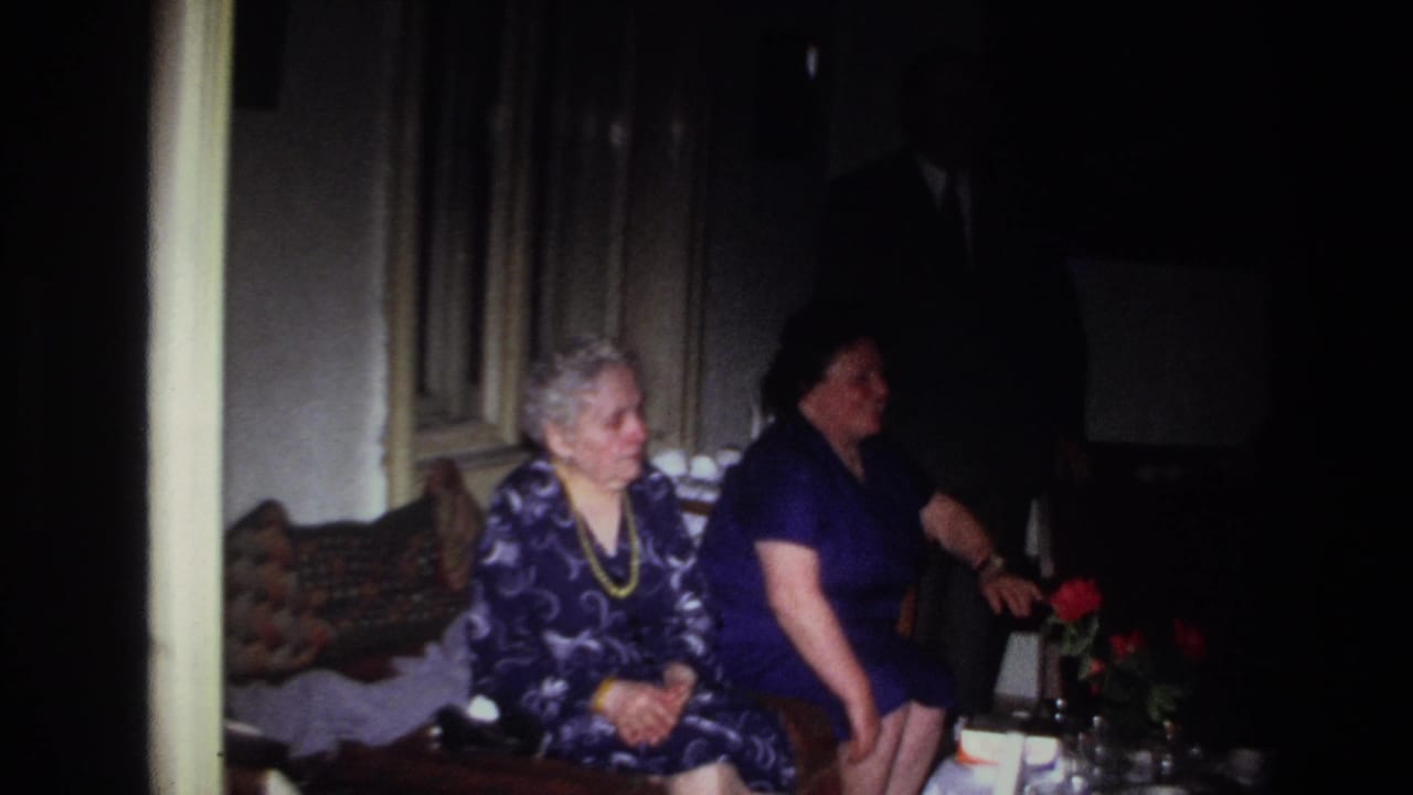 Two seated women are seen as a dark-suited figure stands nearby.
