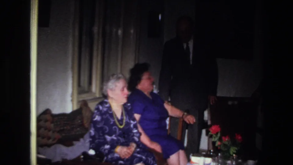 The 1967 France footage shows a man in a dark suit beside two seated women, with red flowers and room furnishings in shadow.