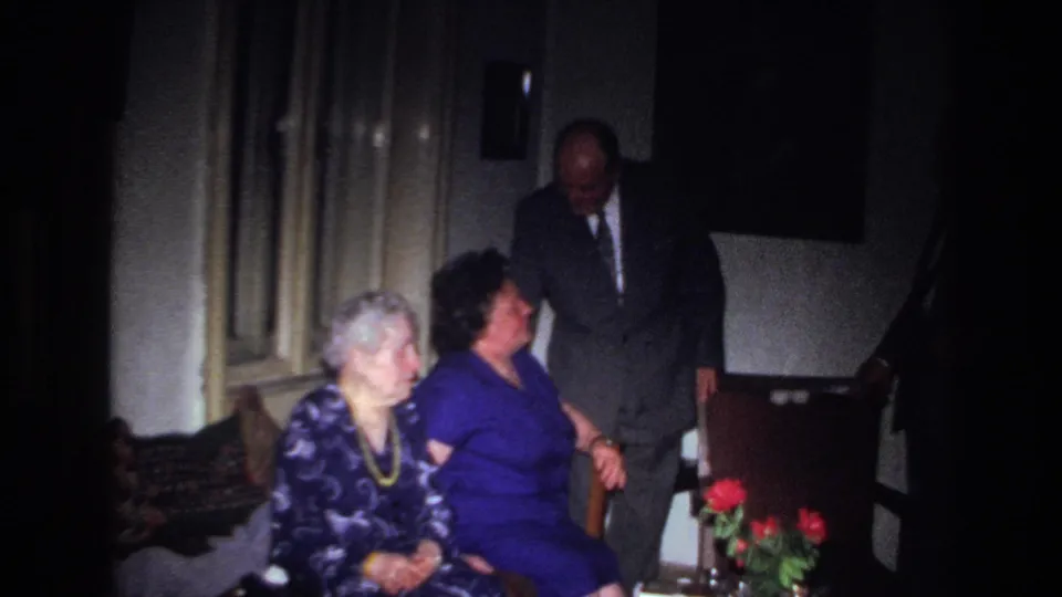 In the dim French interior, a suited man stands smiling near two seated women, with red flowers on a nearby table.